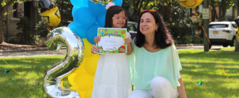 A girl in pink glasses and a white dress standing outside next to a woman in a green shirt who is kneeling next to her. A balloon arch is visible behind them, and the girl is holding a certificate in both hands.