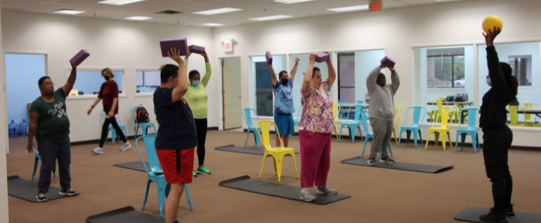 A group of people participating in a yoga class, standing on mats and holding their hands in the air.
