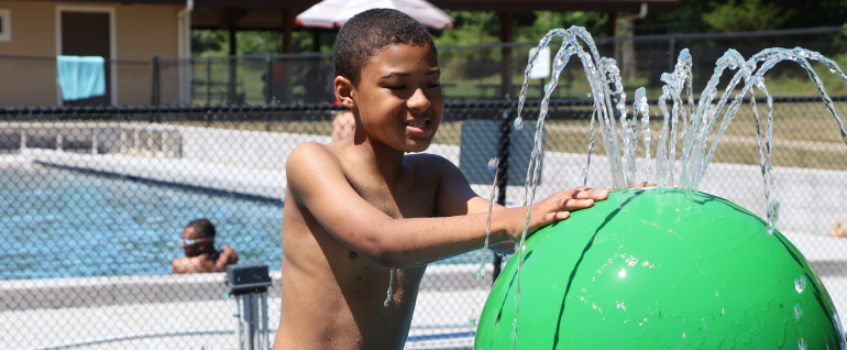 A boy with his hands on a green ball with fountains of water streaming out of the top. A pool is visible in the background.