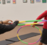 A boy playing with a colorful hula hoop in an indoor activity space; to the left, an instructor holds out a hula hoop.