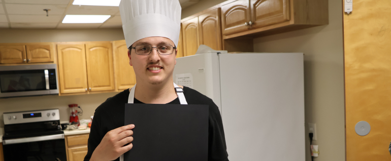 A man standing in a kitchen wearing a black shirt, a white apron. and a white chef's hat. The top of a certificate is visible in his hand.