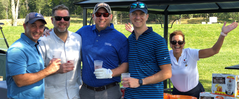 A group of four men on a golf course holding drinks, with a woman behind them at a serving table, with her arms in the air.