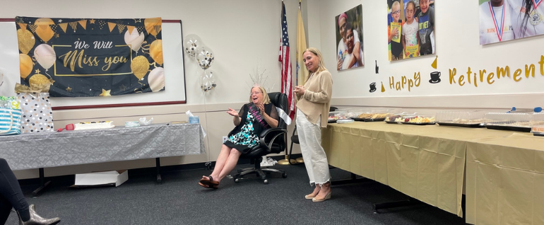 One woman sitting in a chair with another woman standing next to her. All around are decorations that say "We will miss you" and "Happy Retirement."