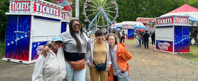 Four women standing together in a carnival, with a Ferris Wheel visible behind them.