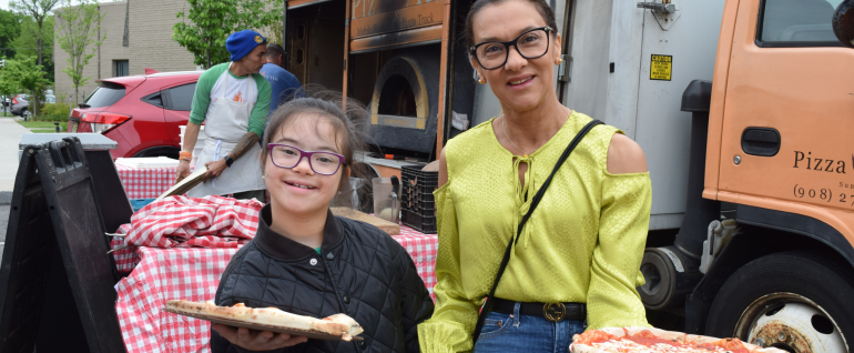 A daughter and mother hold pizza pies in front of a pizza oven food truck. Both are wearing glasses, the girl on the left is in a black shirt, the woman on the right is in a yellow shirt.