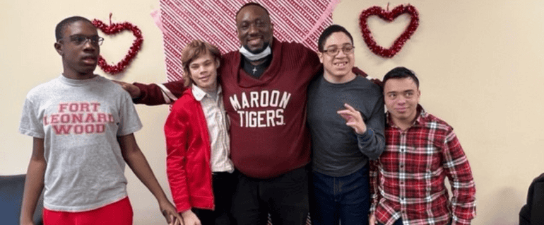Four children gathered on either side of a man in a maroon sweatshirt. Valentine's Day decorations are visible behind them.