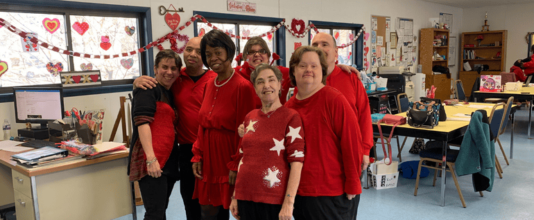 A group of seven adults, all wearing red. Valentines Day decorations are visible in the background.