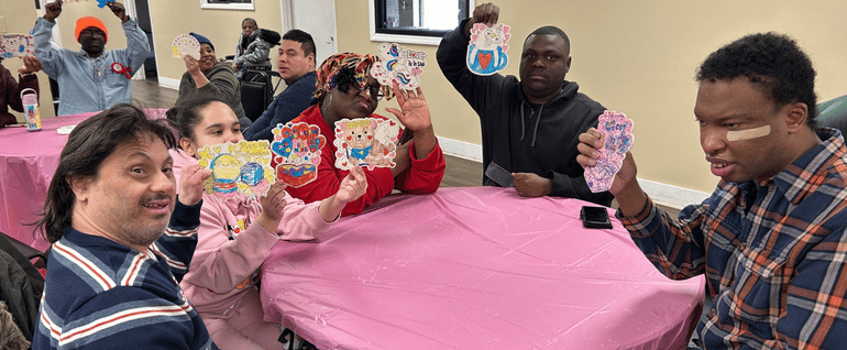 A group of adults gathered around a table with a pink table cloth, with everyone holding up a Valentine's Day craft.