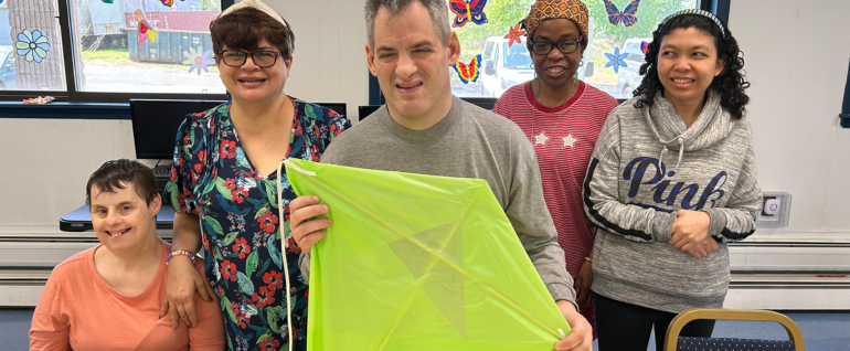 Five adults standing together. The man in the center is holding a bright green kite.