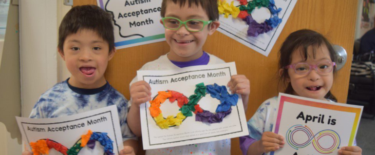 Three children holding papers that have colorful collages on them and say "Autism Acceptance Month."
