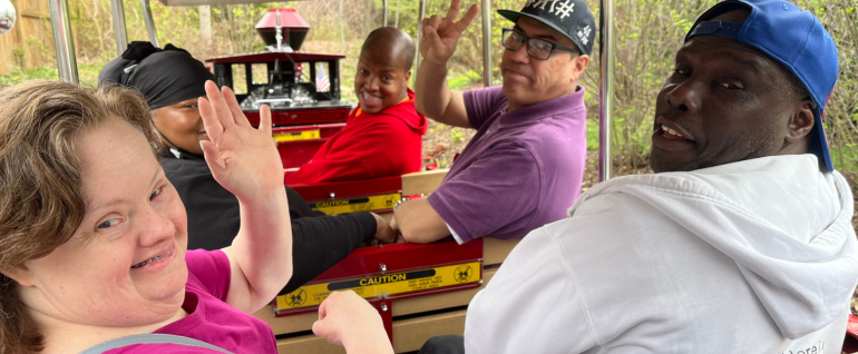 A group of five adults sitting on a train ride, looking back at the camera and waving/smiling.