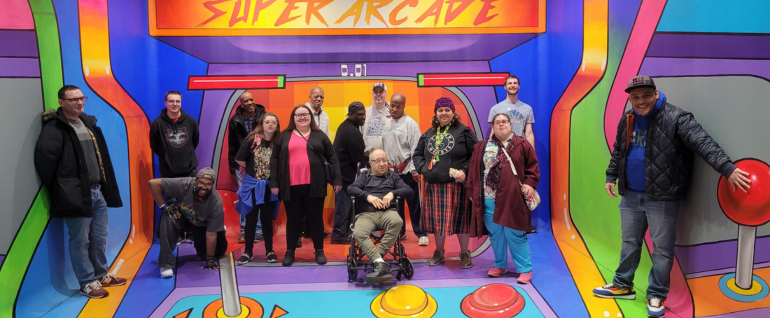 Fifteen adults are standing across a colorful painted scene that looks like the inside of an arcade game, with buttons and joysticks. Across the top are the words "Super Arcade."