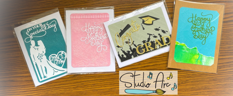 Four cards across a wooden background: Left to right: an "On Your Special Day Mr. & Mrs." card with a turquoise background and white lettering, a pink "Happy Mother's Day" card with white lettering; a gold, brown, and black "Congrats GRAD" card; and a "Happy Father's Day" card with a blue and green background and green lettering. The Studio Arc logo is shown below the cards.