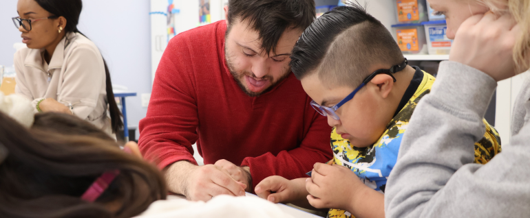 A man in a red shirt coloring alongside a boy with blue glasses and a yellow, blue, and black t-shirt. They are both looking down at the coloring page together. On the far left and far right are school staff. In the background, there is classroom shelving.