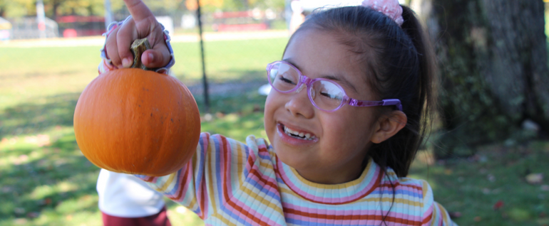 A girl in a striped shirt and pink glasses holds a small pumpkin in one hand.