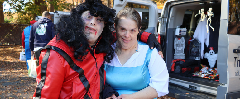 A man dressed in the Michael Jackson Thriller costume and a woman dressed as Dorothy from the Wizard of Oz, with van trunks open behind them with people trick-or-treating.