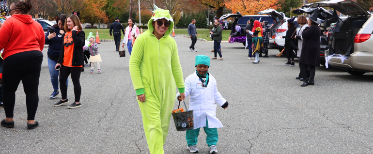 A wide shot of open car trunks for trick or treating, with a teacher wearing a lime green onesie costume holding the hand of a preschooler dressed up as a doctor.