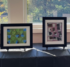 Four pieces of printed art in black frames on a table in front of windows, with fall foliage visible in the background.