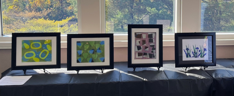 Four pieces of printed art in black frames on a table in front of windows, with fall foliage visible in the background.