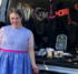 A woman in a dress stands in front of a decorated van with Halloween decor, including a pretend headstone, and candy and treats.