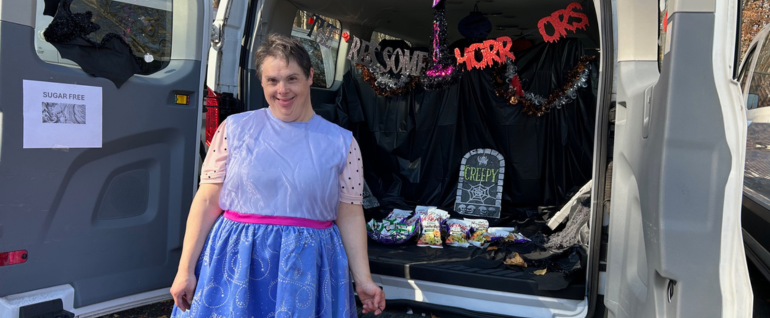 A woman in a dress stands in front of a decorated van with Halloween decor, including a pretend headstone, and candy and treats.