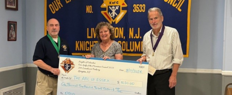 Two men stand on either side of a woman, all holding a giant check made out to The Arc of Essex County. Behind them a banner for the Knights of Columbus Livingston, NJ chapter.