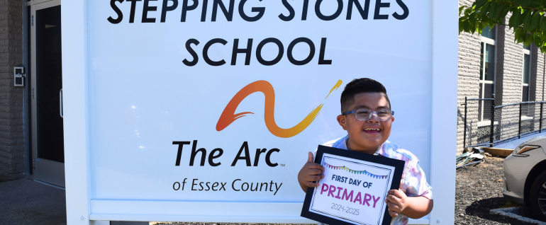 A boy standing in front of a sign for Stepping Stones School holding up a sign that says "First Day of Primary."