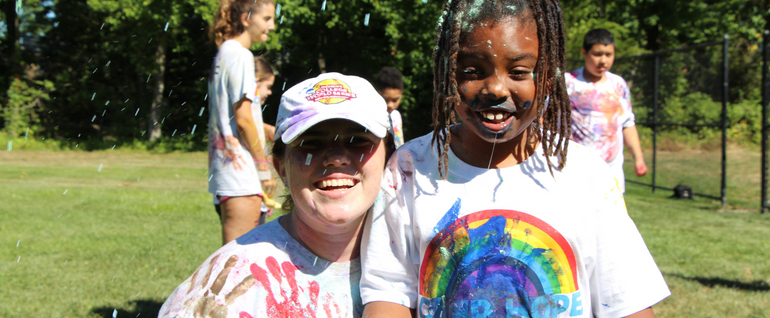 A woman and a child stand together on a lawn wearing Camp Hope t-shirts and covered in paint. Splattering paint is visible in front of them.