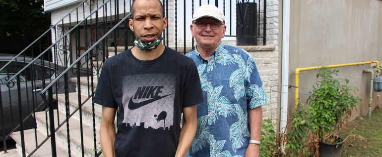 Two men standing near the entrance of a house, with planted herbs visible to the right.
