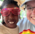 A girl in pink plastic glasses smiles with a woman wearing a Camp Hope t-shirt, under a pavilion at Camp Hope.