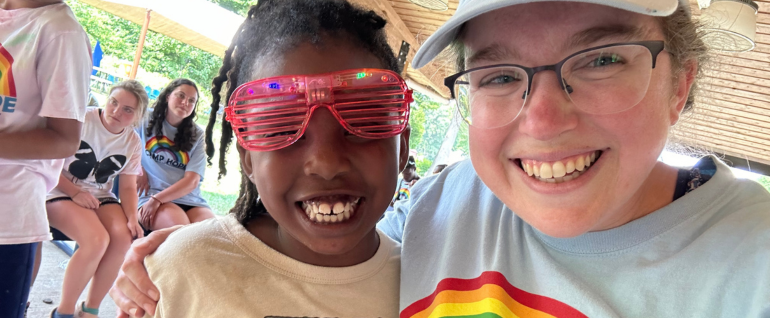 A girl in pink plastic glasses smiles with a woman wearing a Camp Hope t-shirt, under a pavilion at Camp Hope.