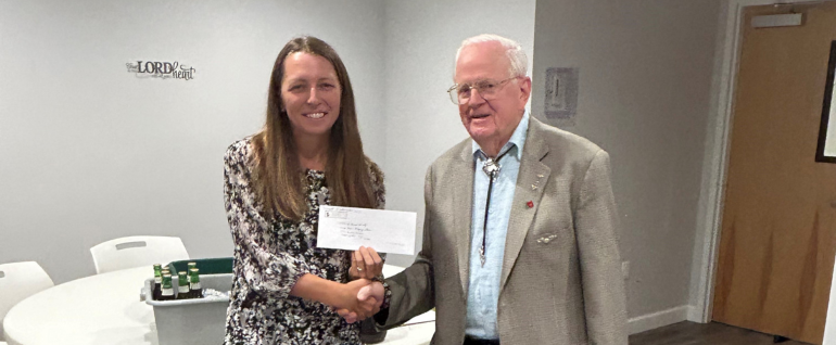 A woman on the left holds a check and shakes hands with a gentleman on the right. Both are smiling at the camera.