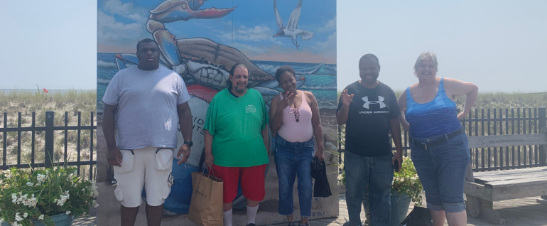 A group of five adults standing on a boardwalk in front of beach dunes, with a mural of the ocean partially behind them.