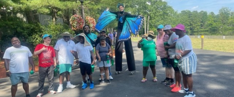 A group of 11 adults standing on a paved path. A woman on stilts with butterfly wings stands among them.