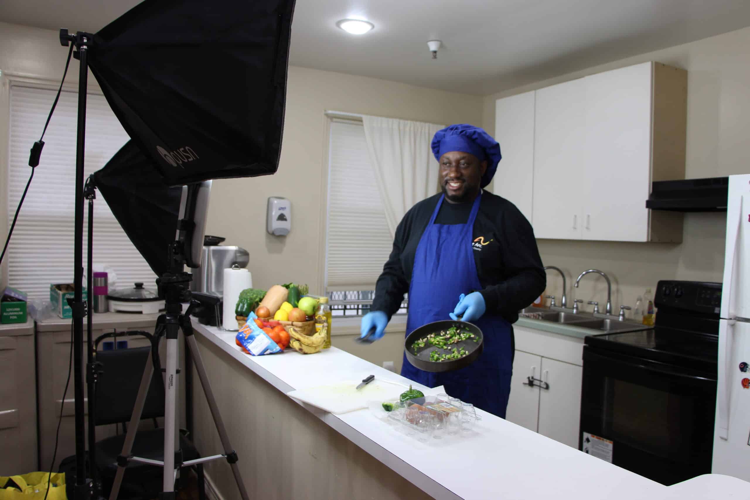 A man wearing a blue chef's hat and apron standing in a kitchen in front of a camera cooking vegetables in a pan.