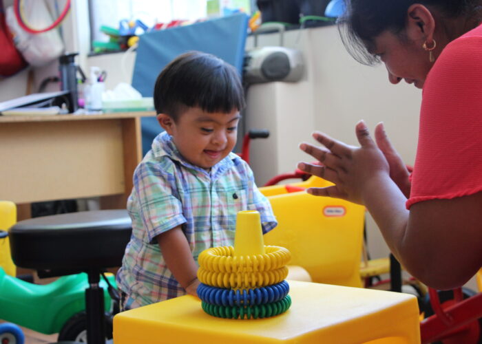 A boy plays with toys while a woman claps her hand. They are in a therapy classroom with colorful plastic chairs and activities.