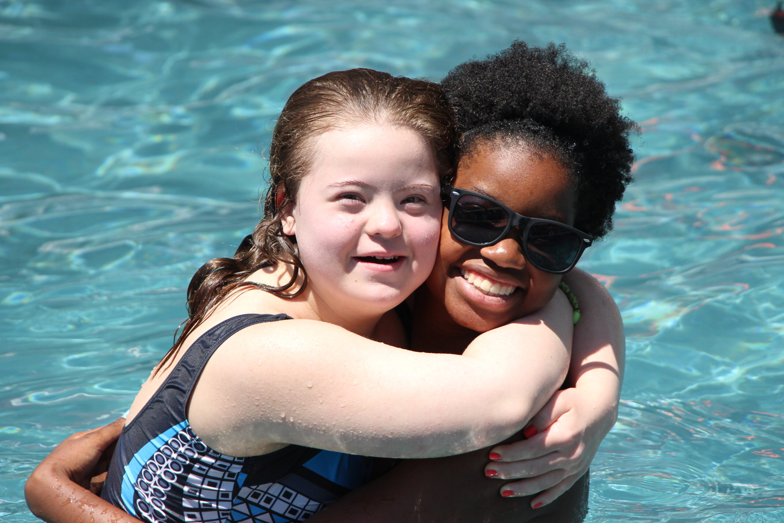 A girl hugs a woman while they are both standing in a swimming pool, smiling at the camera. The woman is wearing sunglasses.