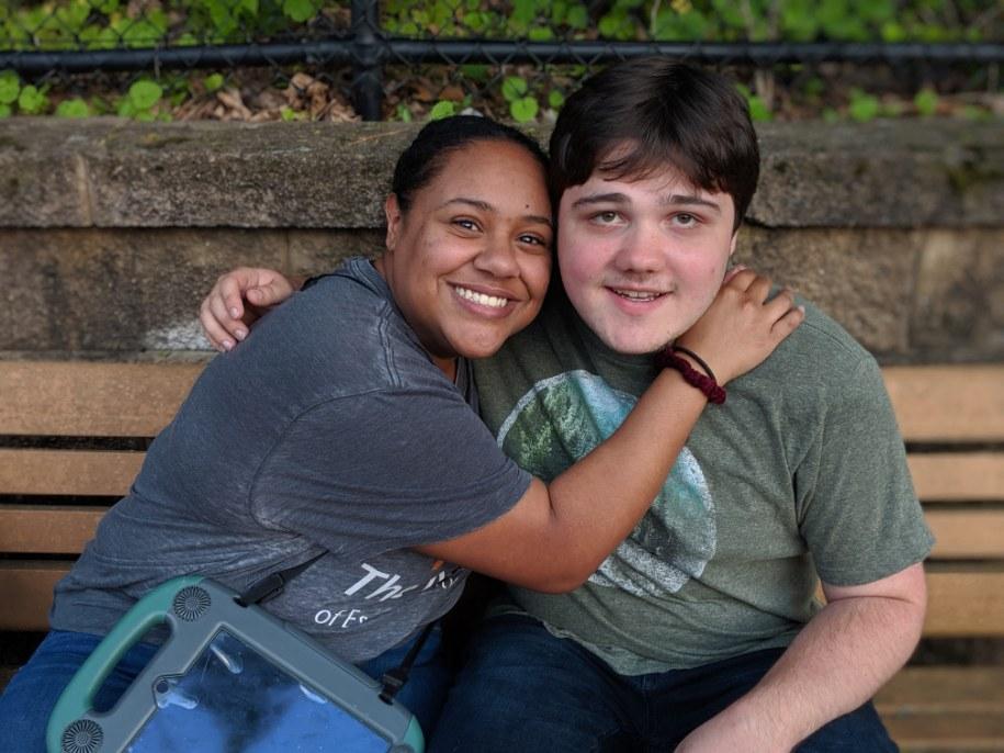 A woman and a teen boy sit together on a park bench and smile at the camera.