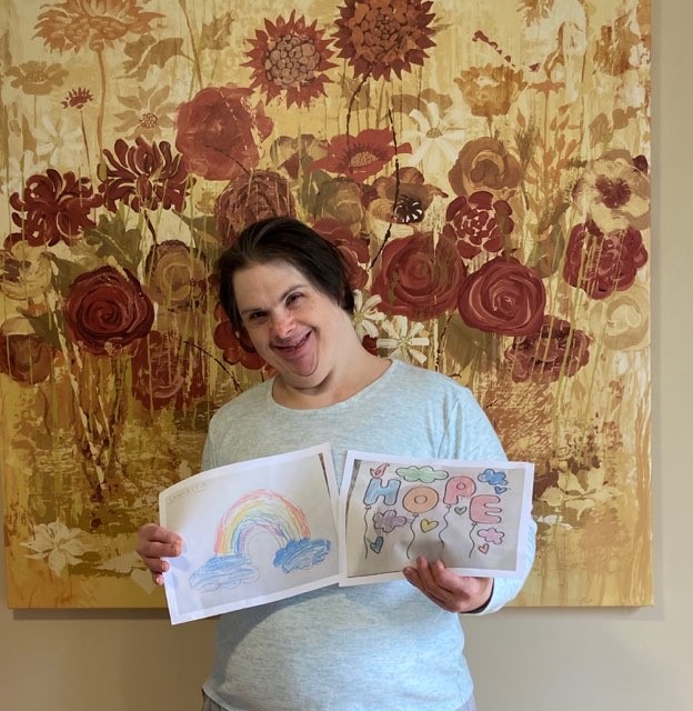 A woman standing in front of a floral painting, holding two drawings: one of a rainbow, and one with the word 