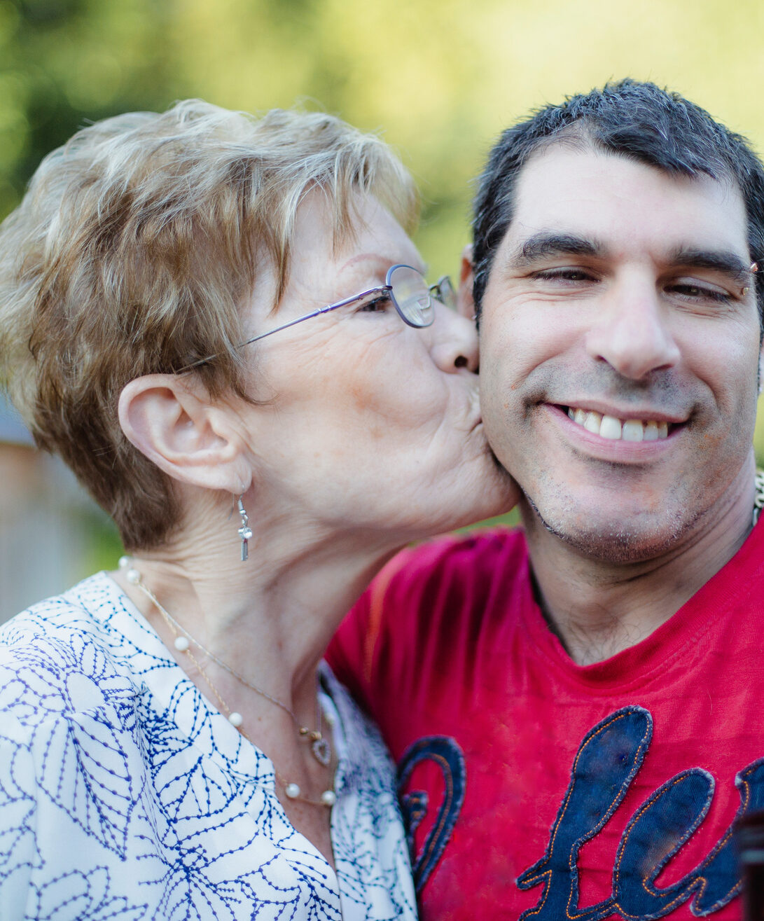A woman in glasses and a white and blue shirt kissing the cheek of a young man in a red shirt.
