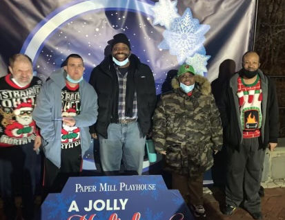 A group of people in holiday attire standing in front of a snow globe backdrop and in front of a sign that says "Paper Mill Playhouse: A Jolly Holiday."