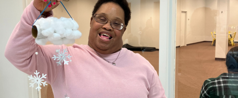 A woman in a pink sweater holds up a craft of a cotton-ball cloud with snowflakes underneath.