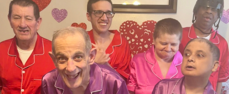 A group of six adults wearing red, purple, and pink pajamas, with Valentine's Day decorations on the wall behind them.