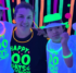 Three children and a teacher smile together in a black-light party, with fluorescent hats and necklaces and shirts that say "Happy 100 Days of School"