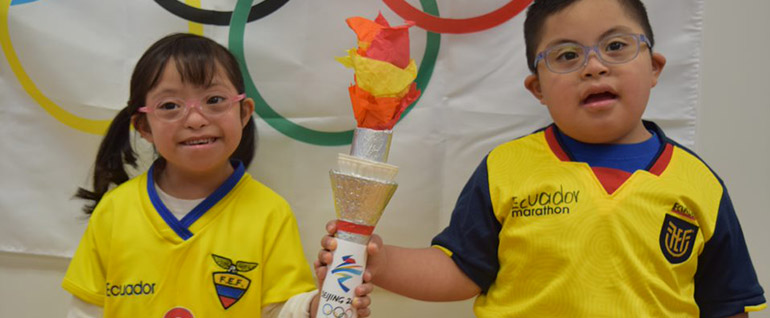 Two children wearing Ecuador soccer jerseys and holding an Olympic torch made with foil and tissue paper. The Olympic rings are visible behind them.