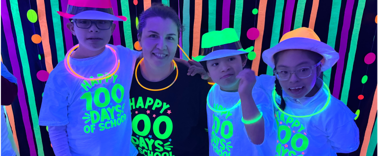 Three children and a teacher smile together in a black-light party, with fluorescent hats and necklaces and shirts that say "Happy 100 Days of School"