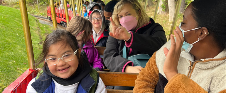 Students and staff sitting on a train ride outdoors. The child in the front seat is smiling and wearing glasses.