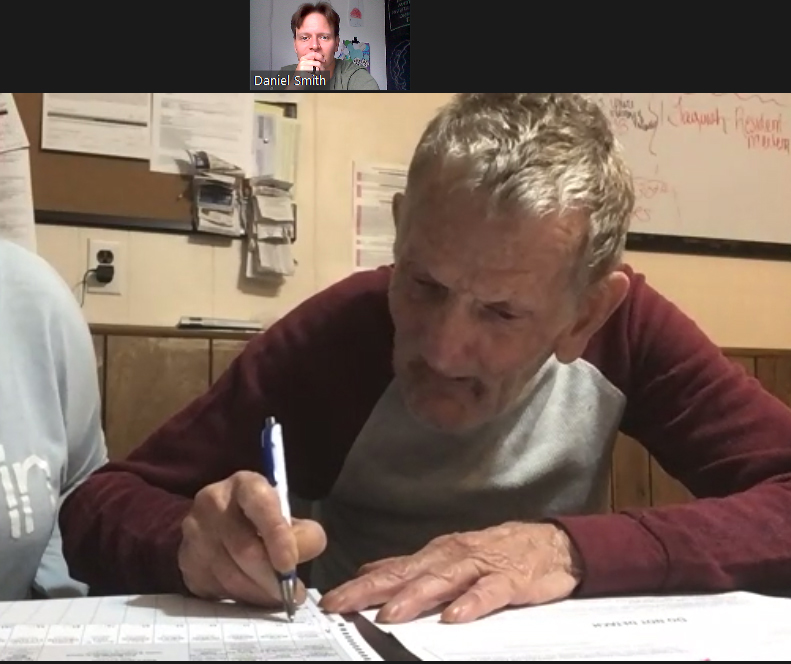 A screenshot of man filling out a mail-in ballot at a table with an inset screen of a man watching.