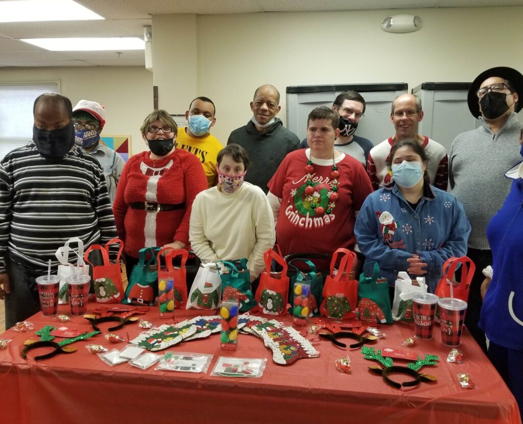 A group of 11 people wearing festive holiday attire and standing behind a table filled with holiday gift bags and decorations.