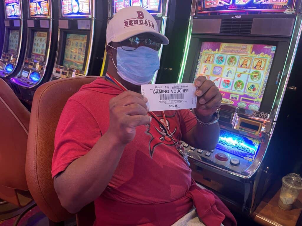 A man in a red shirt and a blue mask sits at a slot machine and holds a gaming voucher in his hand.